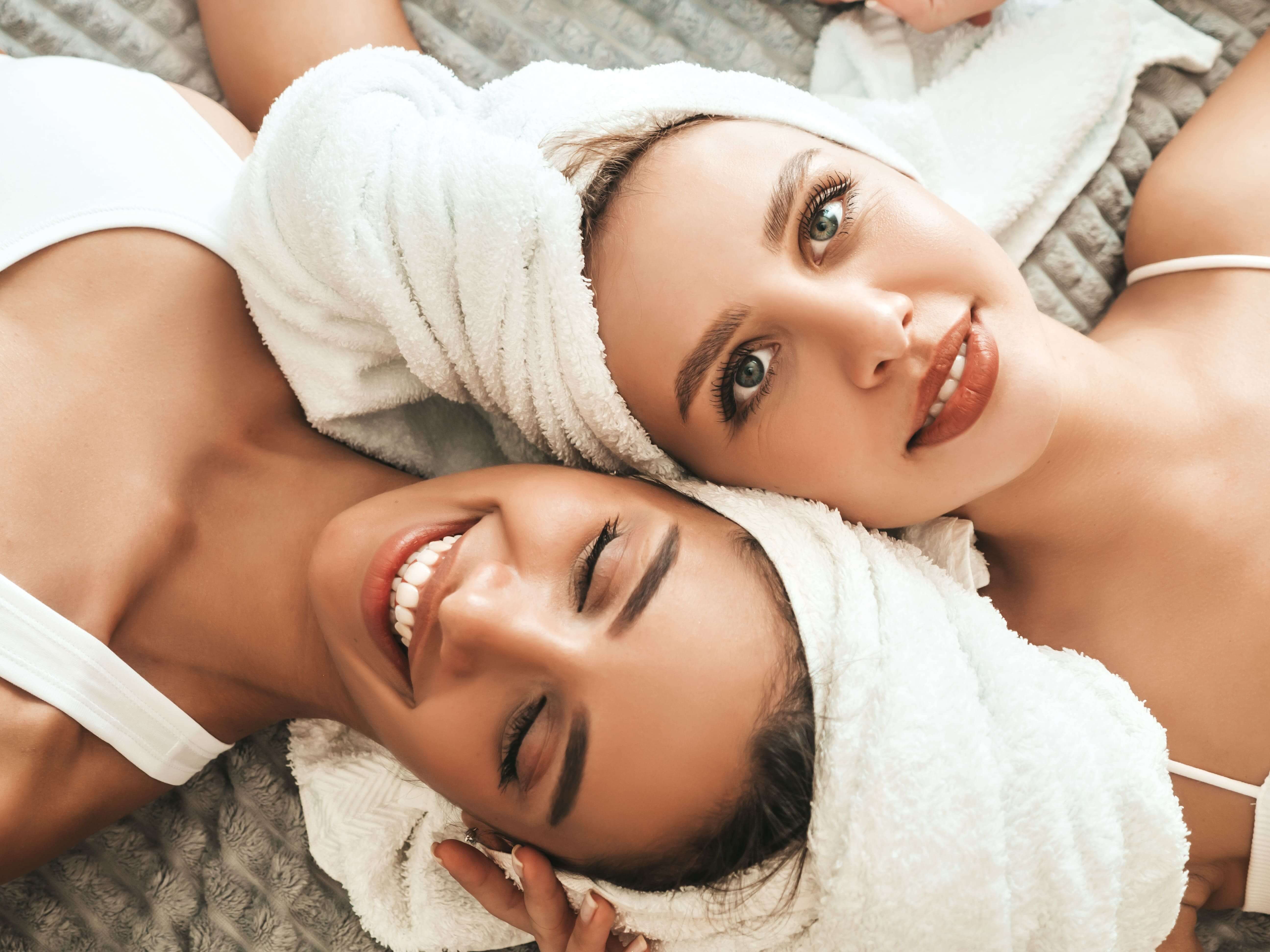 two young beautiful smiling women white bathrobes towels head min