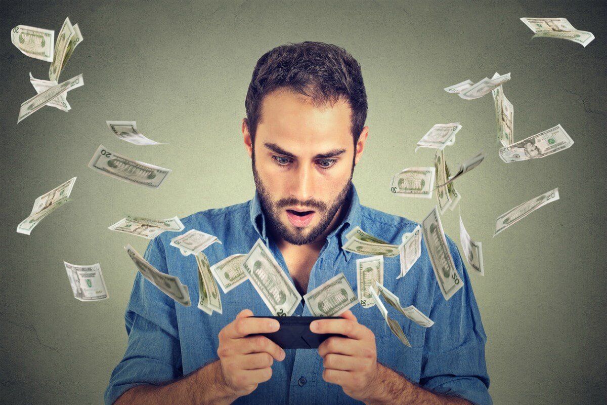 Technology online banking money transfer, e-commerce concept. Shocked young man using smartphone with dollar bills flying away from screen isolated on gray wall office background.